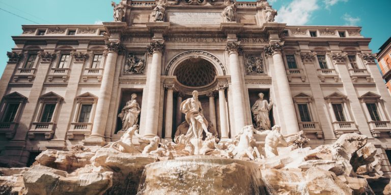 Das Bild zeigt den berühmten Trevi-Brunnen (Fontana di Trevi) in Rom, Italien. Im Vordergrund befindet sich die prachtvolle Brunnenanlage mit eindrucksvollen Skulpturen, darunter der Meeresgott Oceanus in der Mitte, flankiert von Tritonen und Pferden. Im Hintergrund steht die reich verzierte barocke Fassade des Palazzo Poli. Der Himmel ist klar und blau, was dem Bild eine warme, einladende Atmosphäre verleiht.